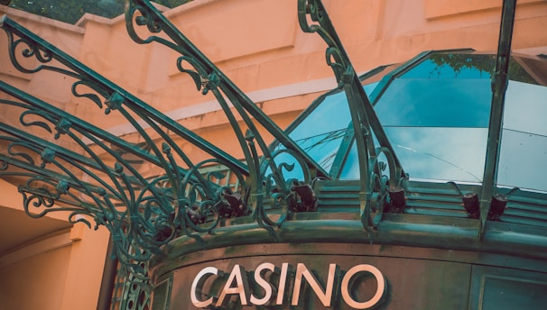 The image displays the entrance to a casino, characterized by detailed and ornate architectural elements with green metal structures and a sign that reads 'CASINO'. The background includes a glass dome and a peach-colored wall.